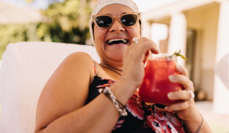 A joyful woman in sunglasses, holding a red drink, laughs while reclining outdoors.