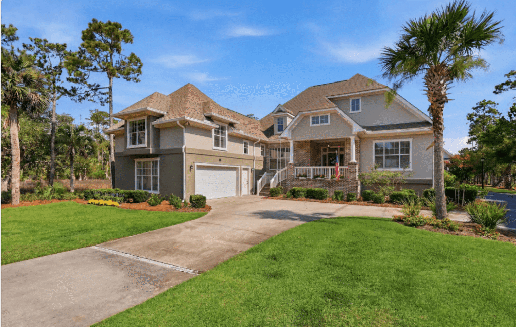 Two-story suburban house with three-car garage, front porch, and well-kept lawn, surrounded by palm trees and landscaping on a sunny day.