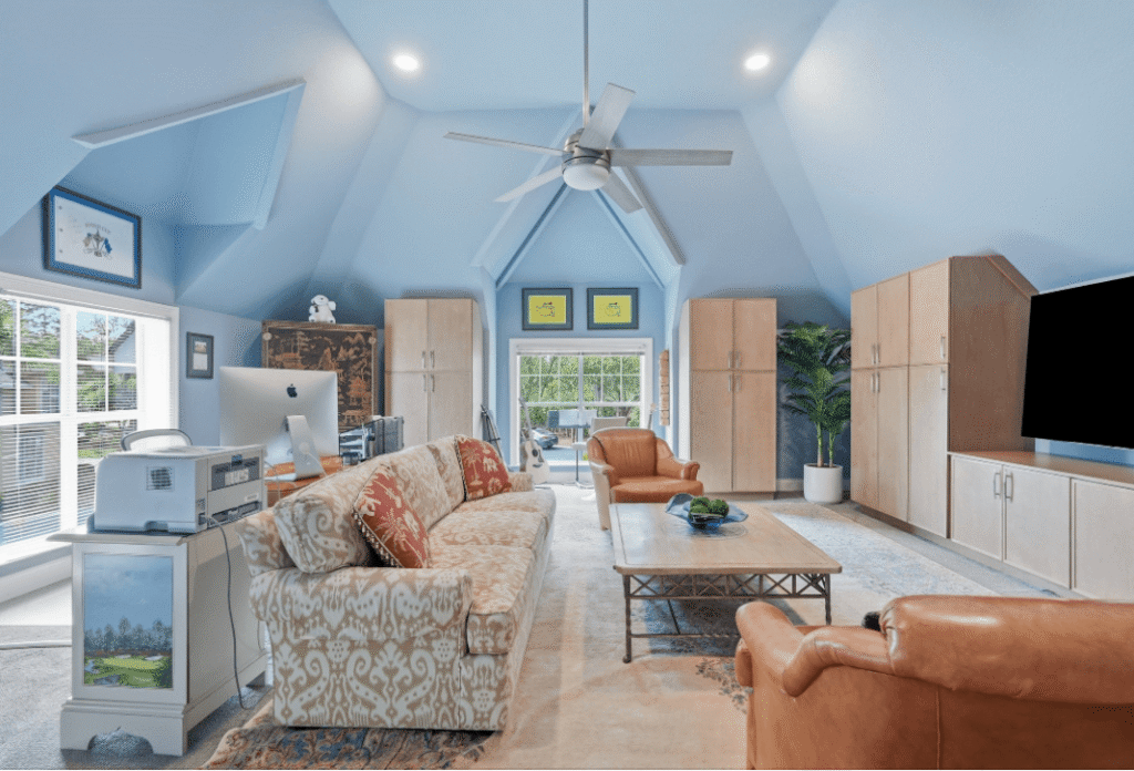 Spacious attic living room with blue walls, beige couches, a large TV, computer desk, ceiling fan, and windows letting in natural light.