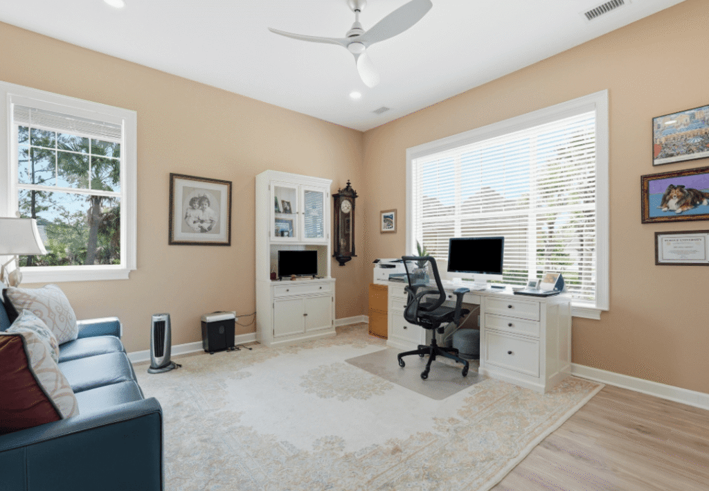 Home office with two desks, computers, a blue sofa, framed art, a large rug, and big windows letting in natural light; neutral walls and wood flooring complete the room.