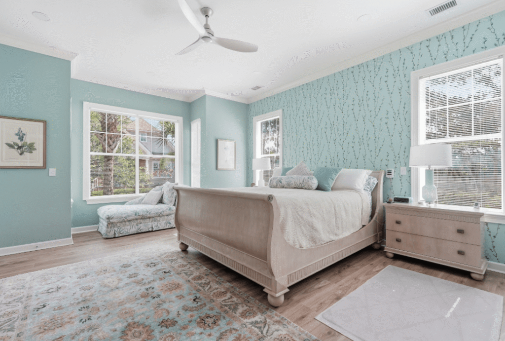 Bright bedroom with a wooden bed, patterned blue accent wall, large windows, chaise lounge, rug, and nightstand with lamp.