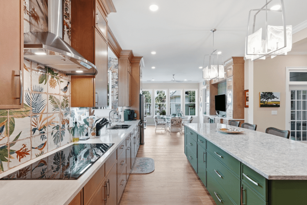 Bright kitchen with wood cabinets, green island, marble countertops, patterned tile backsplash, modern lighting, and an open view to a living area with windows and glass doors leading outside.