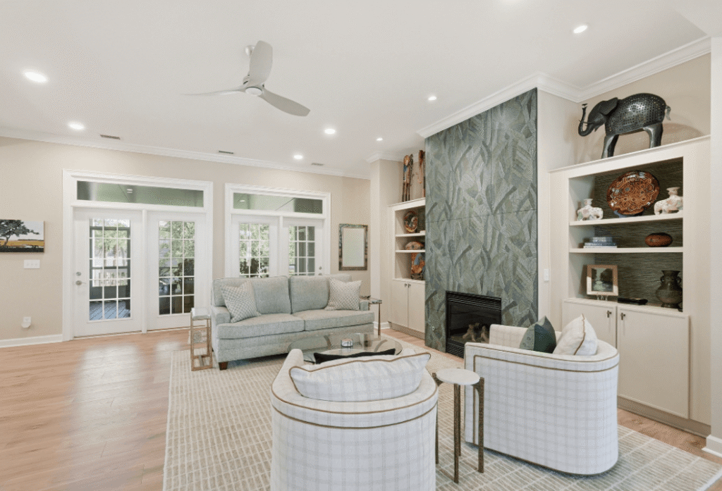 Modern living room with patterned accent wall, fireplace, gray sofa, two white chairs, built-in shelves, hardwood floors, and glass doors leading to an outdoor area.