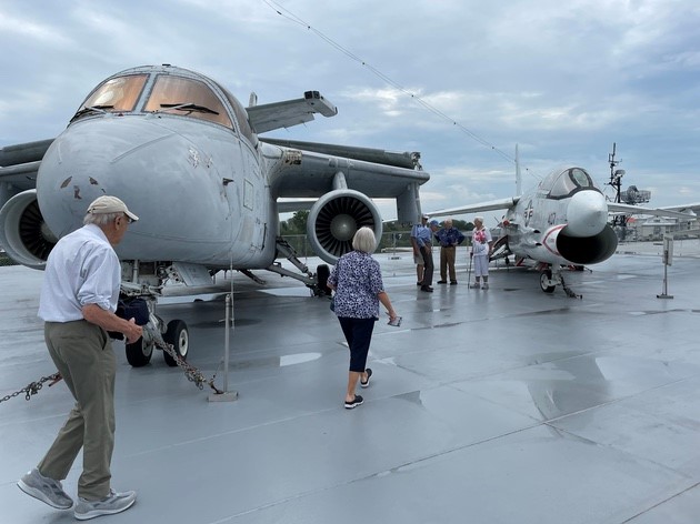A group of people looking at a group of fighter jets on the deck of a ship.