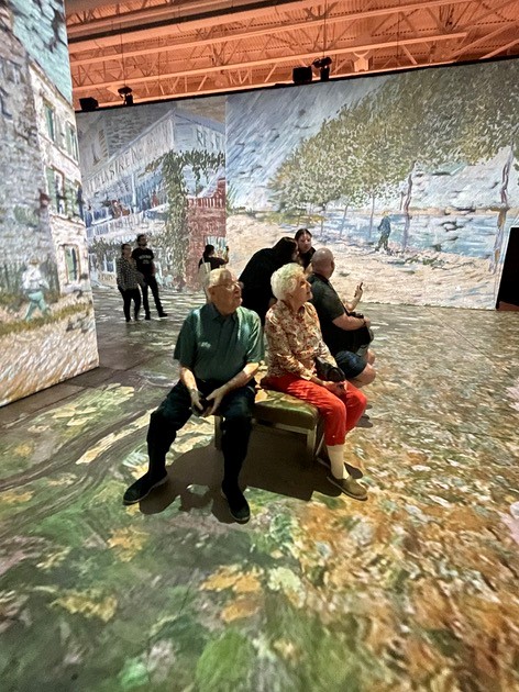A group of people sitting on a bench in front of a large painting.