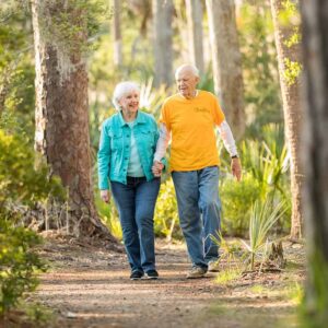 An elderly couple holding hands while walking on a forest trail, surrounded by tall pine trees and sunlight filtering through.